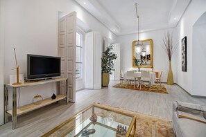 Un salon moderne doté d’un téléviseur à écran plat, d’une table basse en verre, d’un canapé et d’un coin repas avec une table et des chaises blanches.