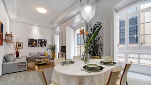 A dining area with a round table set for four, surrounded by chairs, with a sofa and a television in the background.