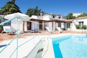 A pool area with lounge chairs, a covered seating area, and a building with a tiled roof.