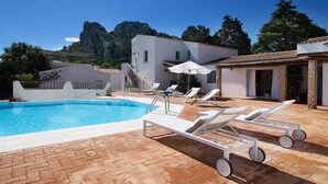 Una piscina con sillas de descanso y sombrillas, un edificio con tejado de azulejos y montañas al fondo.