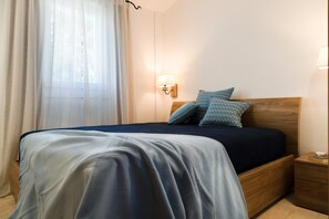 A bedroom with a bed, a nightstand, a wall-mounted lamp, and sheer curtains.