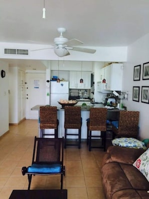 A compact living space with a kitchenette, a dining area with three wicker chairs, a sofa, and a ceiling fan.