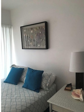 A bedroom with a bed, two pillows, a nightstand with a lamp, a framed artwork, and a window with curtains.