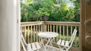 Un balcon avec une table blanche et deux chaises blanches, un pot de fleurs avec des fleurs roses, et une vue sur un feuillage vert et luxuriant.
