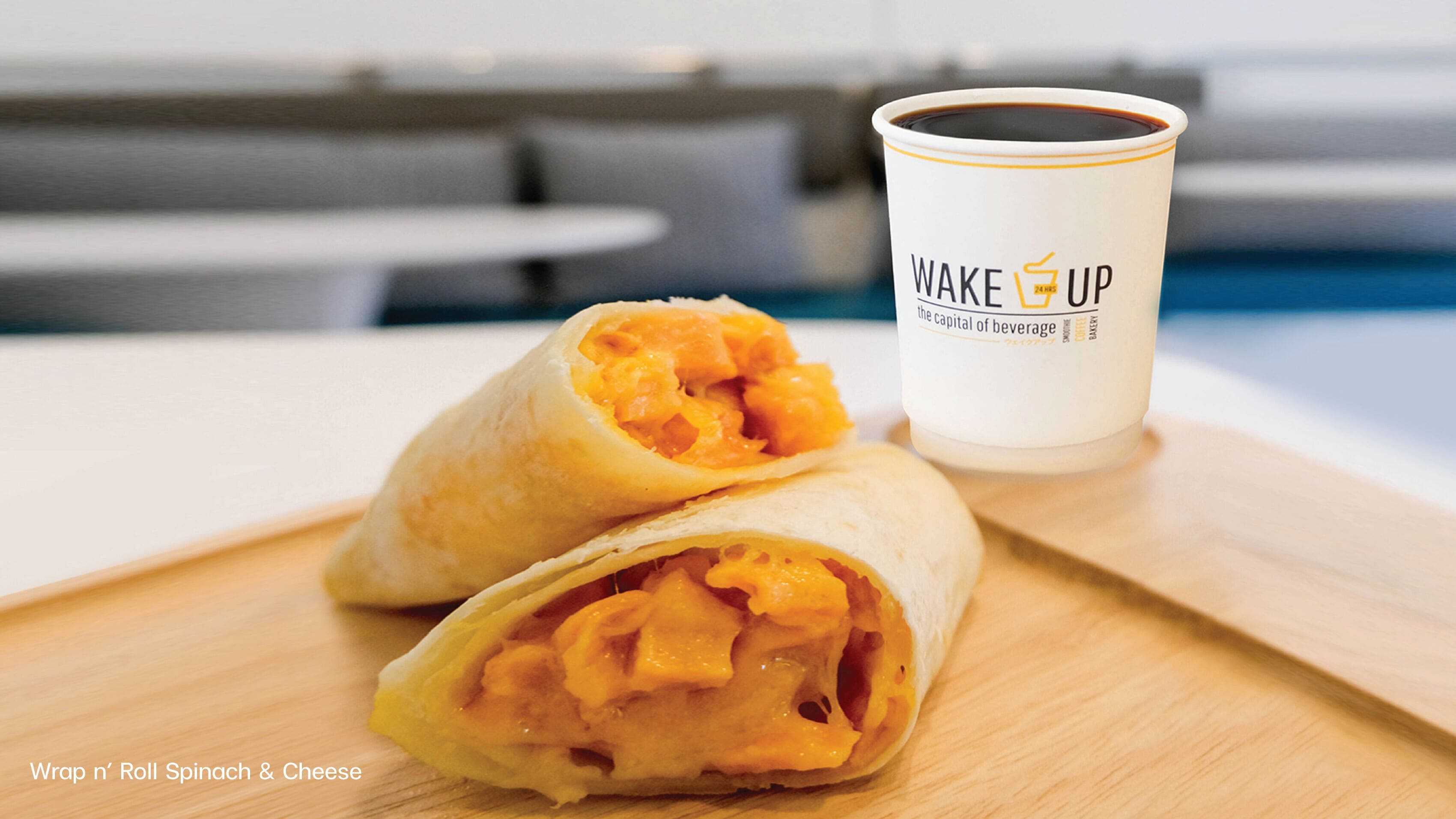 Wrap met spinazie en kaas op een houten plank, naast een kop koffie met de tekst 'Wake Up' en 'de hoofdstad van de drankjes' op een tafel.