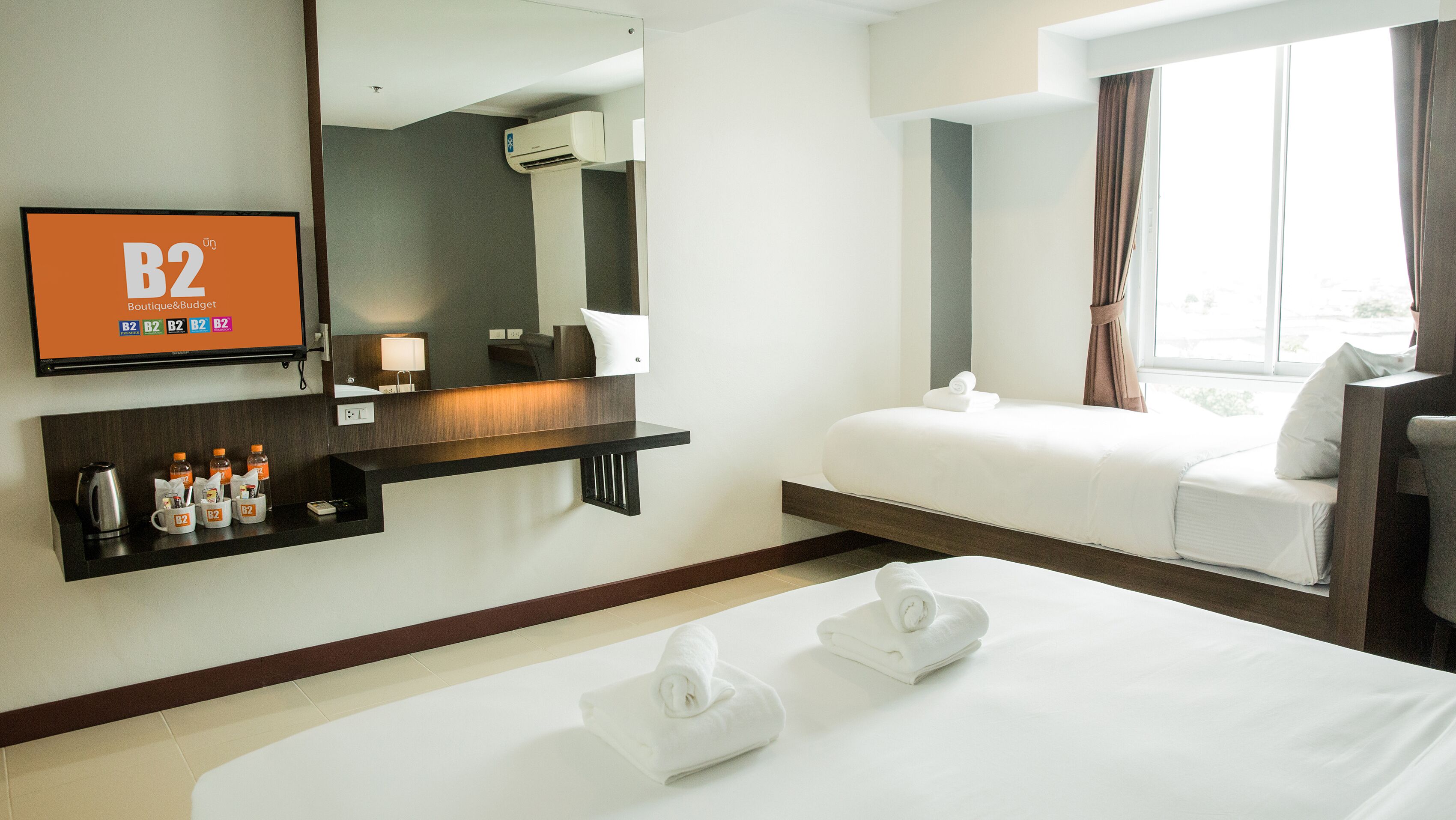 A hotel room with a bed, a bedside table with a lamp, a TV displaying 'B2 Boutique & Budget', and a window with curtains.