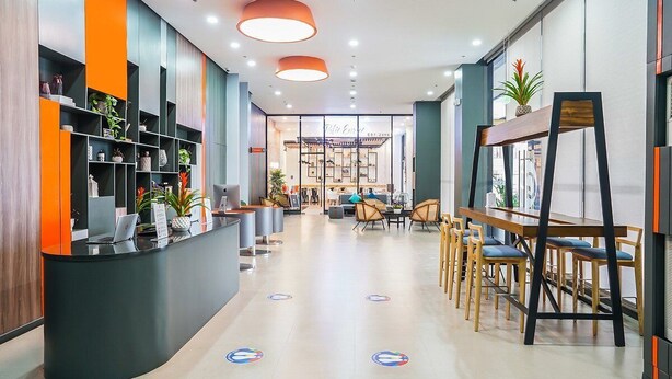 A modern reception area with a curved black reception desk, shelves with plants, and a seating area with wooden chairs and tables.