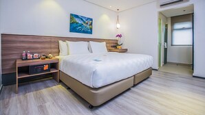 A modern hotel room with a large bed, a bedside table with a digital clock, a small shelf with toiletries, and a painting on the wall.