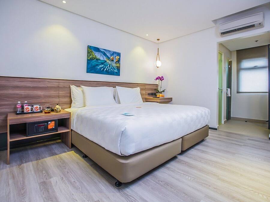 A modern hotel room with a large bed, a bedside table with a digital clock, a small shelf with toiletries, and a painting on the wall.