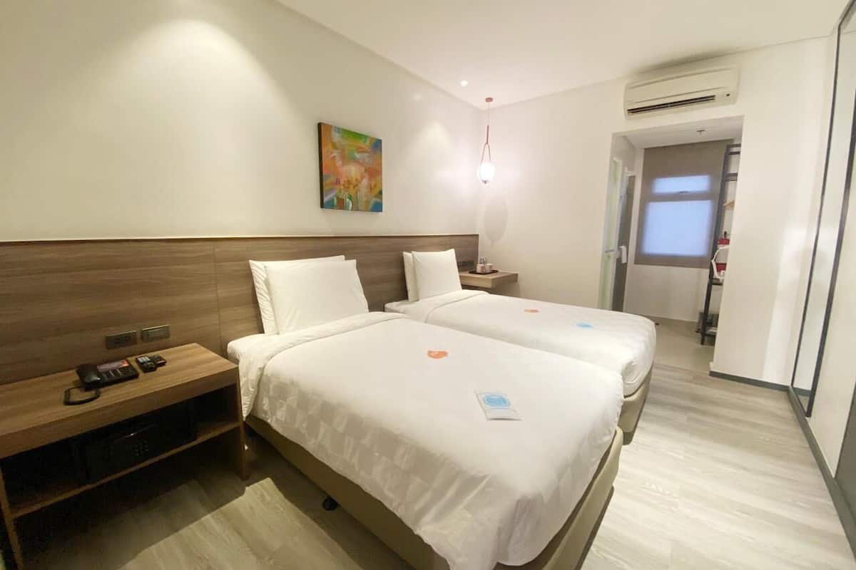 Habitación de hotel con dos camas, cabecero de madera, mesita de noche con teléfono, un cuadro en la pared y un equipo de aire acondicionado.