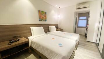 A hotel room with two beds, a wooden headboard, a bedside table with a phone, a painting on the wall, and an air conditioning unit.