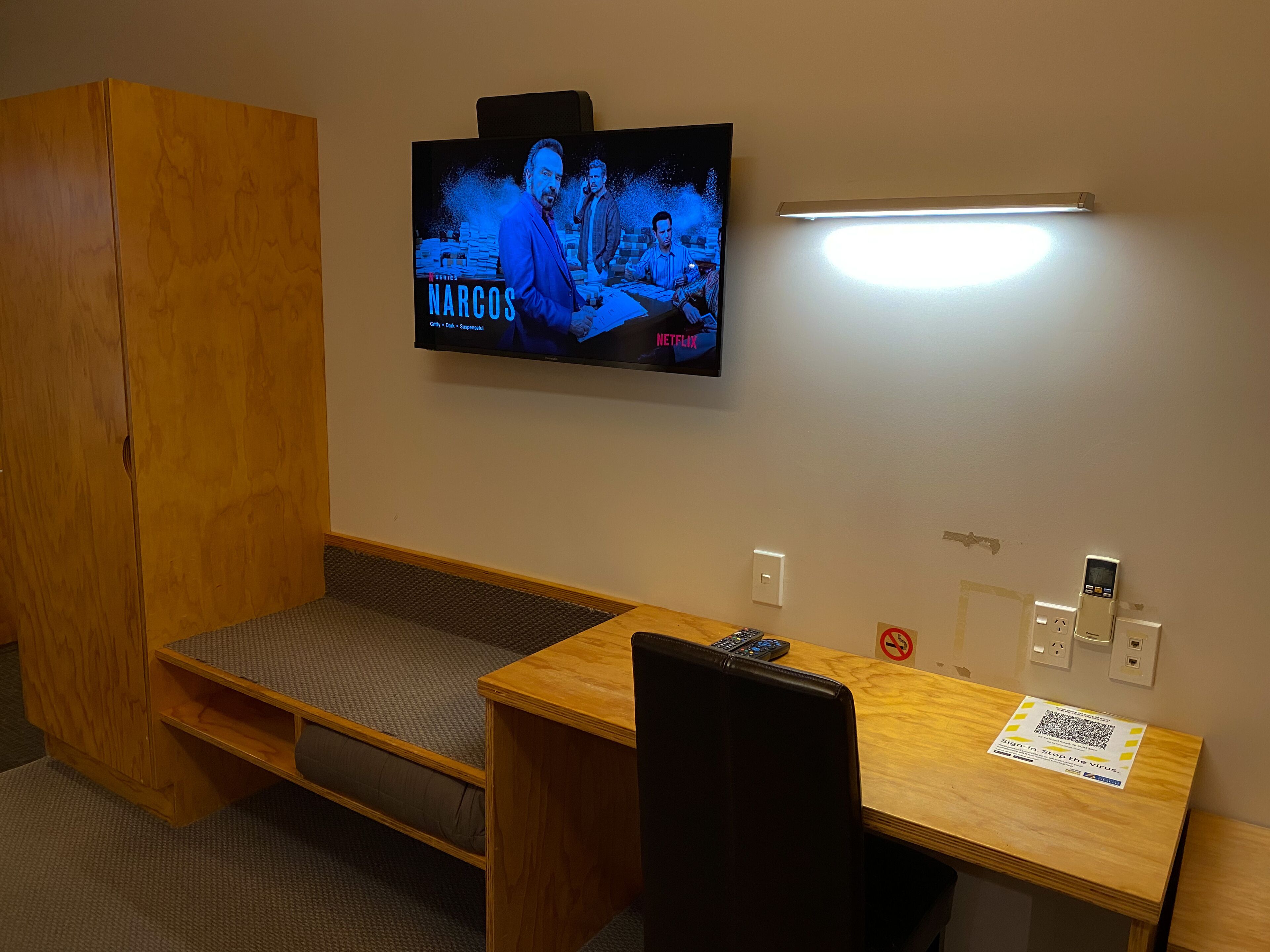 A small room with a wooden desk, a bench, a TV mounted on the wall, and a sign on the wall.