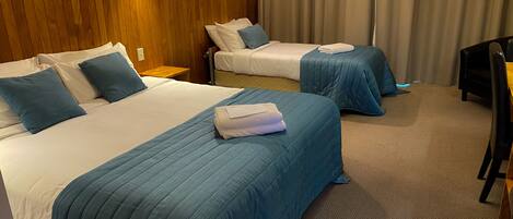 A hotel room with two beds, each with blue bedding and pillows, a wooden headboard, a small wooden nightstand, and a chair.
