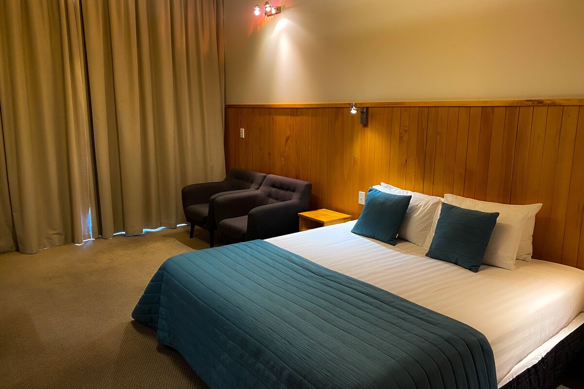 A hotel room with a bed, a chair, a nightstand, and a wooden headboard.