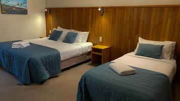 A hotel room with two beds, each with blue bedding and white pillows. There is a wooden headboard, a small wooden nightstand, and a painting on the wall.