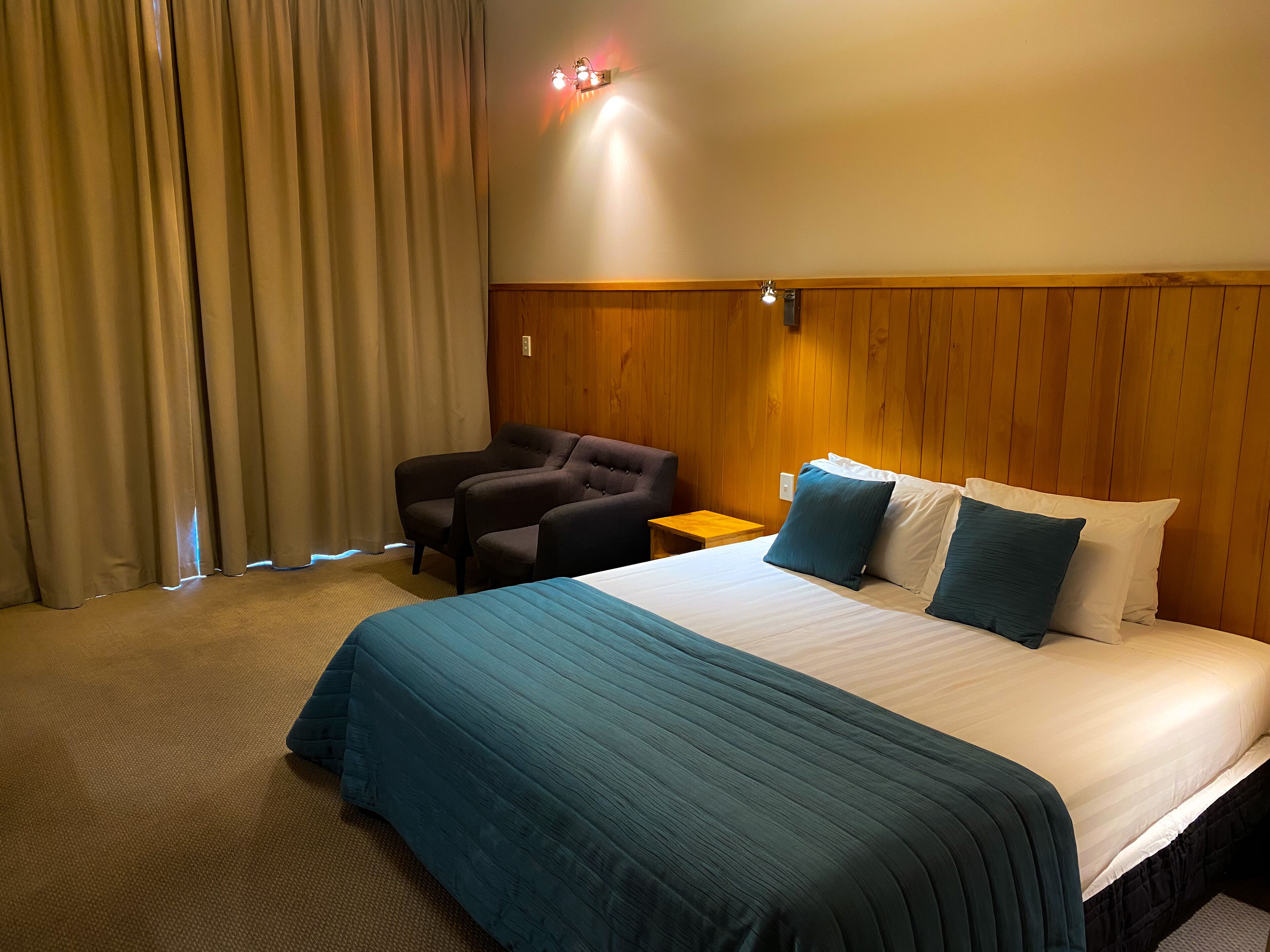 A hotel room with a large bed, a wooden headboard, a nightstand, a chair, and a window with curtains.