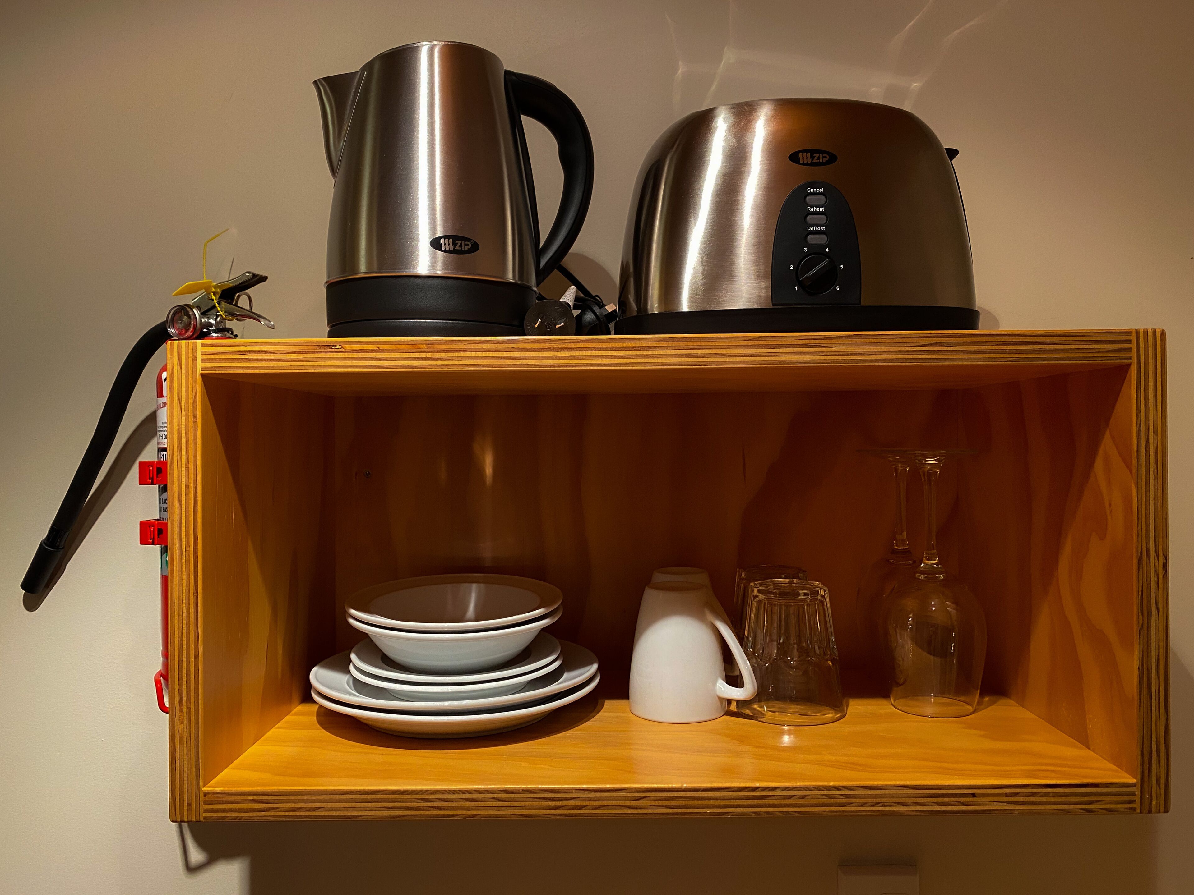 Una estantería de madera con una tetera, una cafetera, platos apilados, una jarra y un vaso.