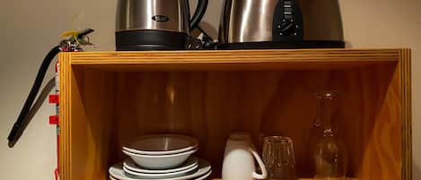 A wooden shelf with a kettle, a coffee maker, stacked plates, a pitcher, and a glass.