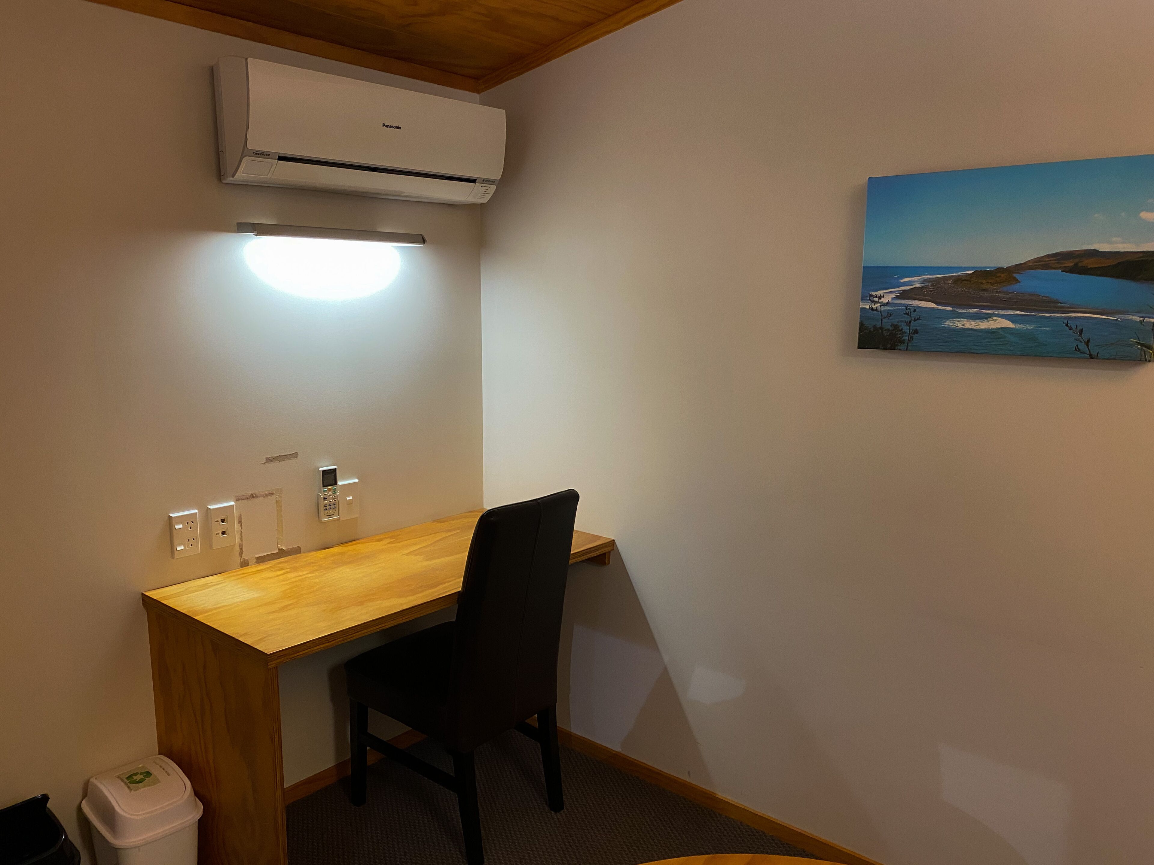 Una habitación pequeña con un escritorio de madera, una silla, un aire acondicionado, un enchufe y un cuadro de un paisaje costero.