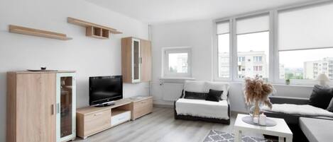 A modern living room with a TV, sofa, and a view of buildings through large windows.
