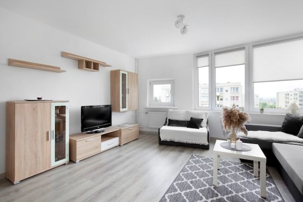 A modern living room with a TV, sofa, and a view of buildings through large windows.