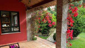 A covered patio with a red wall, a wooden table, a potted plant, and a view of a garden with red flowers. - WATER FRONT SECRET GARDEN (ORTIGUEIRA)