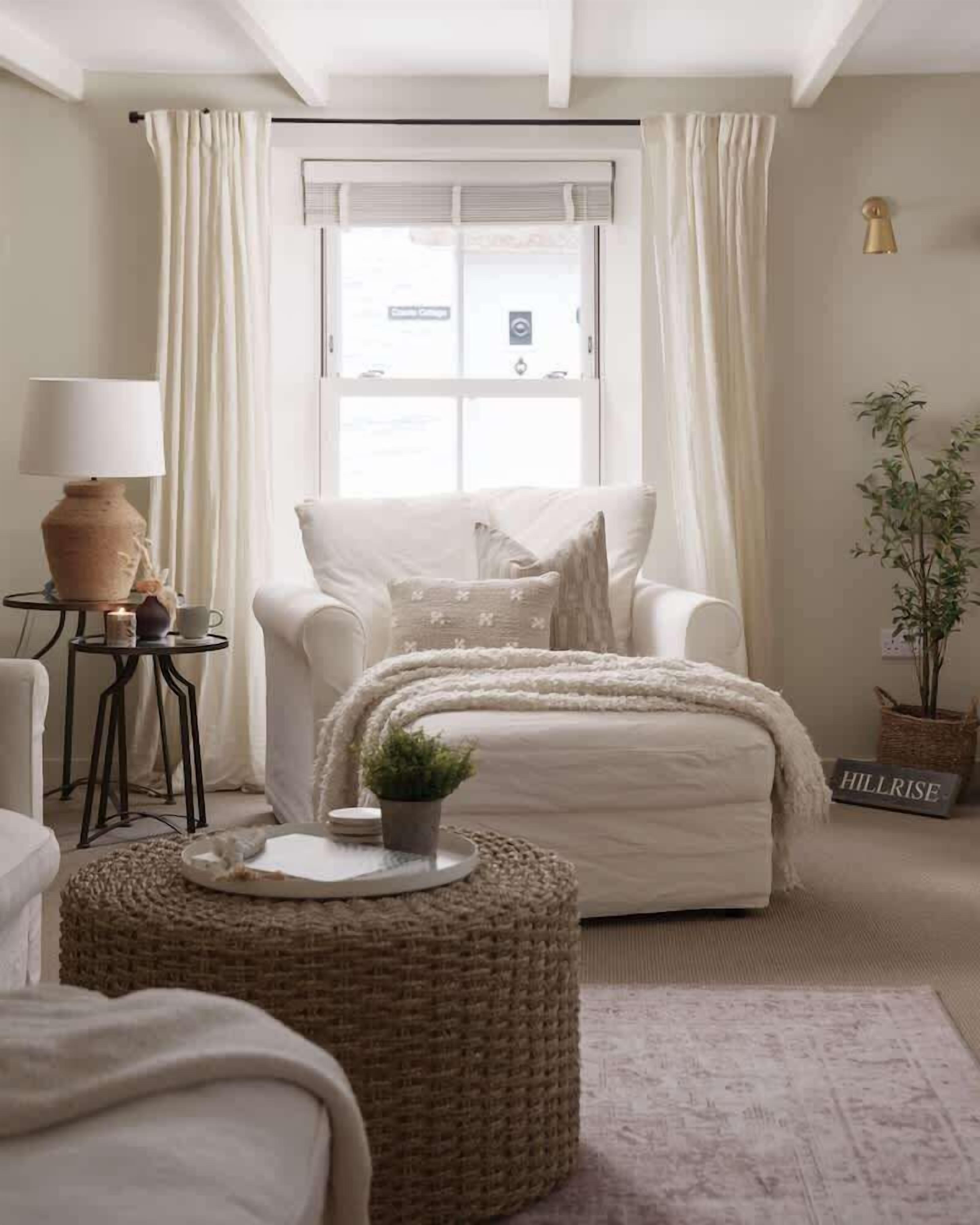 Une pièce chaleureuse avec une grande fenêtre, un fauteuil blanc, une petite table avec une lampe et une plante en pot.