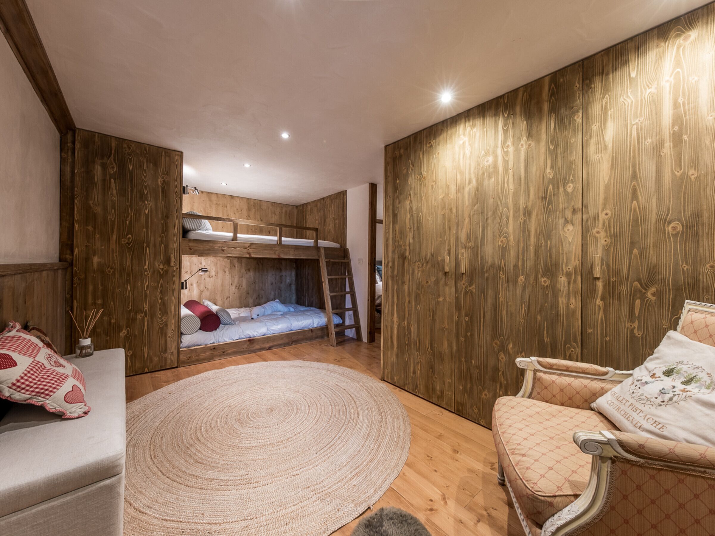 A cozy room with a bunk bed, a round rug, a cushioned chair, and wooden paneling.