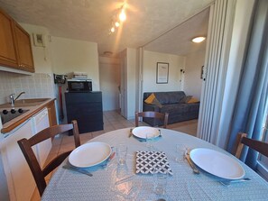 Een compacte keuken met een magnetron, spoelbak en fornuis, een eettafel gedekt met borden en glazen, en een grijze bank in de woonkamer.