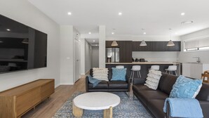 Een moderne woonkamer met een grote televisie, een zithoek, een salontafel en een keuken op de achtergrond.