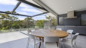 Un espacio moderno para comer al aire libre con una mesa redonda de madera y sillas blancas, con vistas a un paisaje de árboles y cielo despejado.