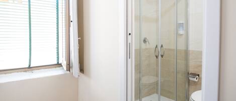 A bathroom with a glass-enclosed shower, a toilet, and a window with blinds.