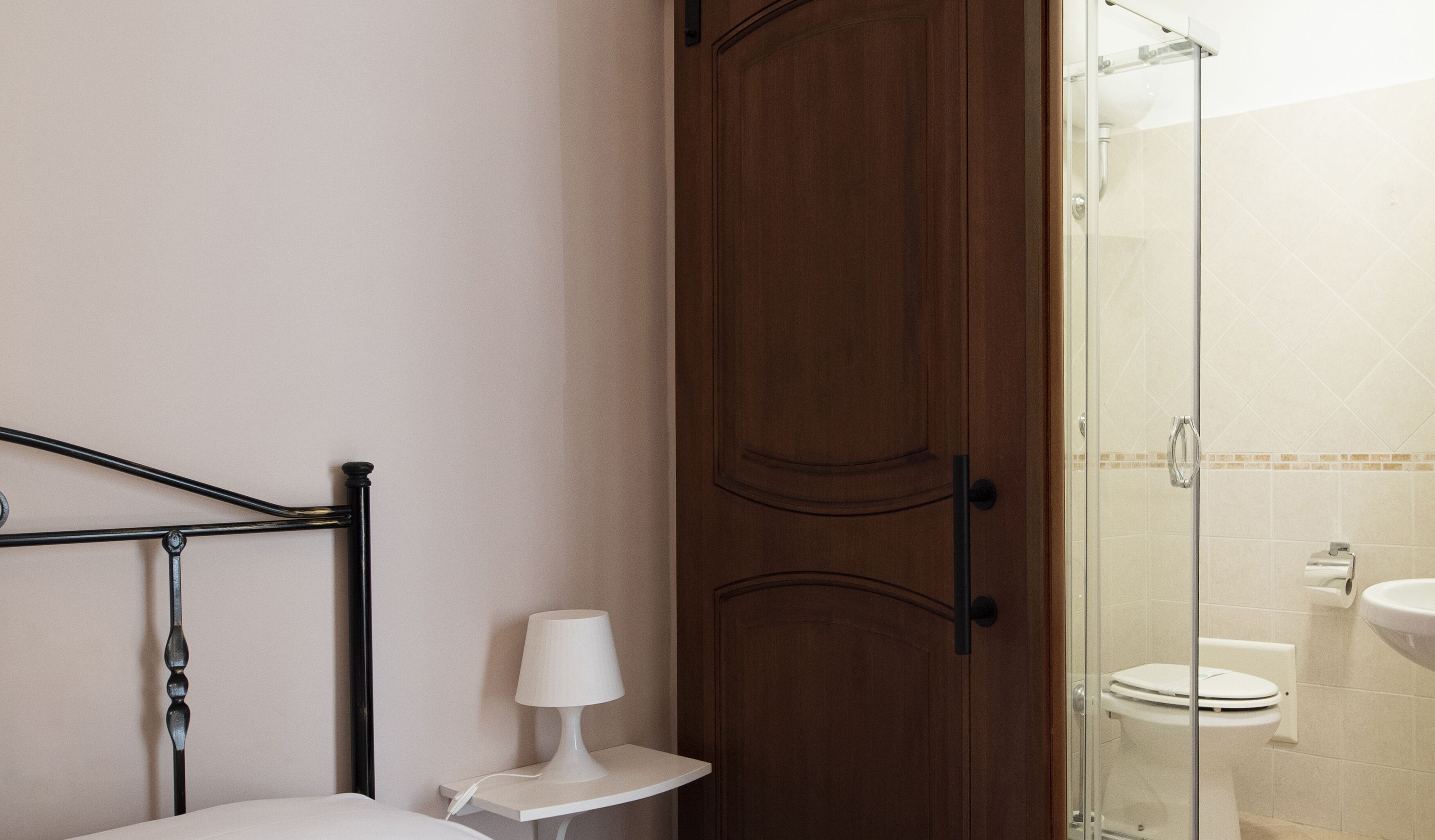 Un baño con puerta de madera, cabina de ducha de vidrio, inodoro y mesita de noche con lámpara.