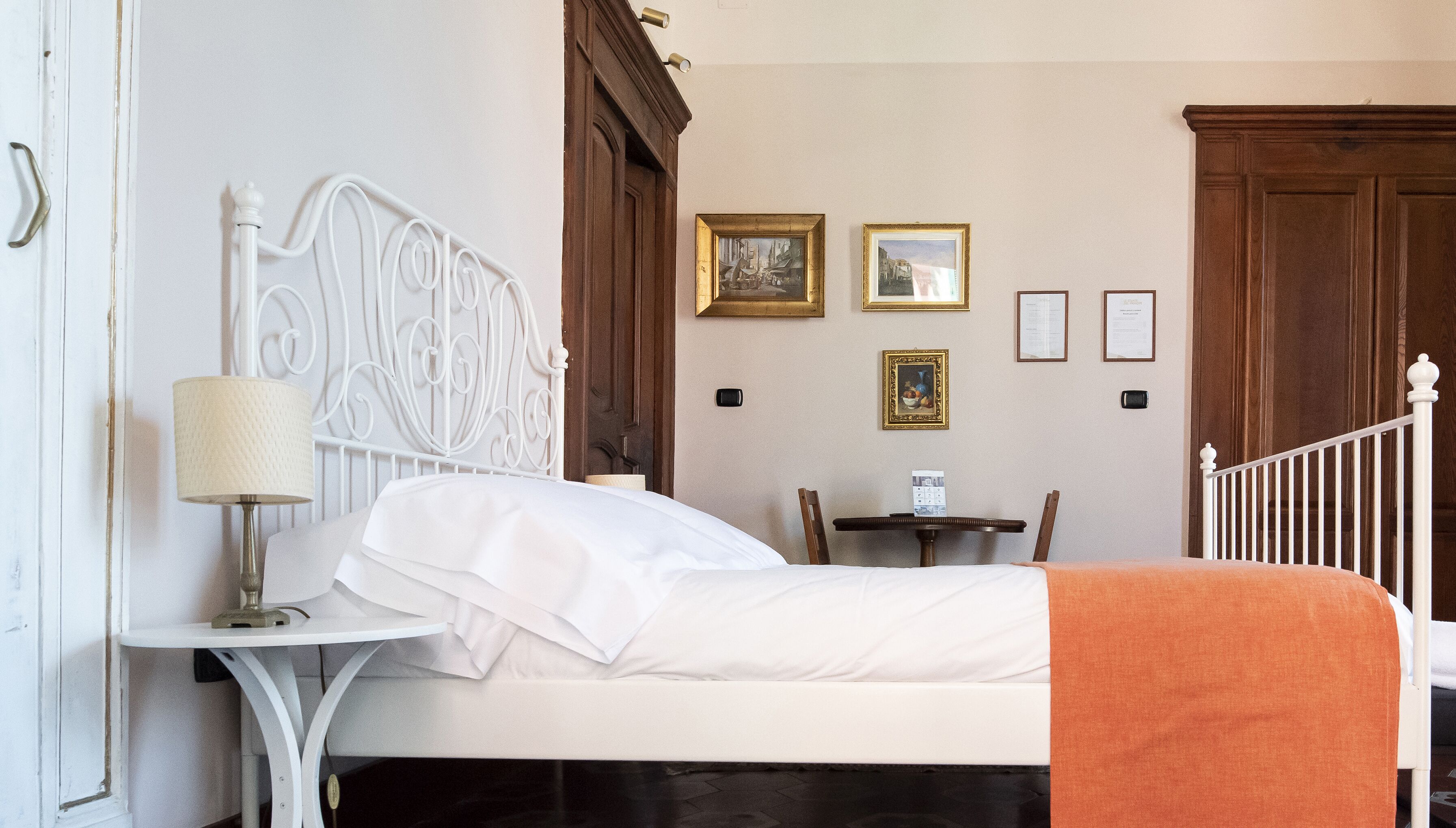 Una cama bien tendida con sábanas blancas y un plaid naranja, cabecera de metal blanco, una mesita de noche con lámpara y una silla de madera.