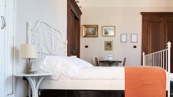 Una cama bien tendida con sábanas blancas y un plaid naranja, cabecera de metal blanco, una mesita de noche con lámpara y una silla de madera.