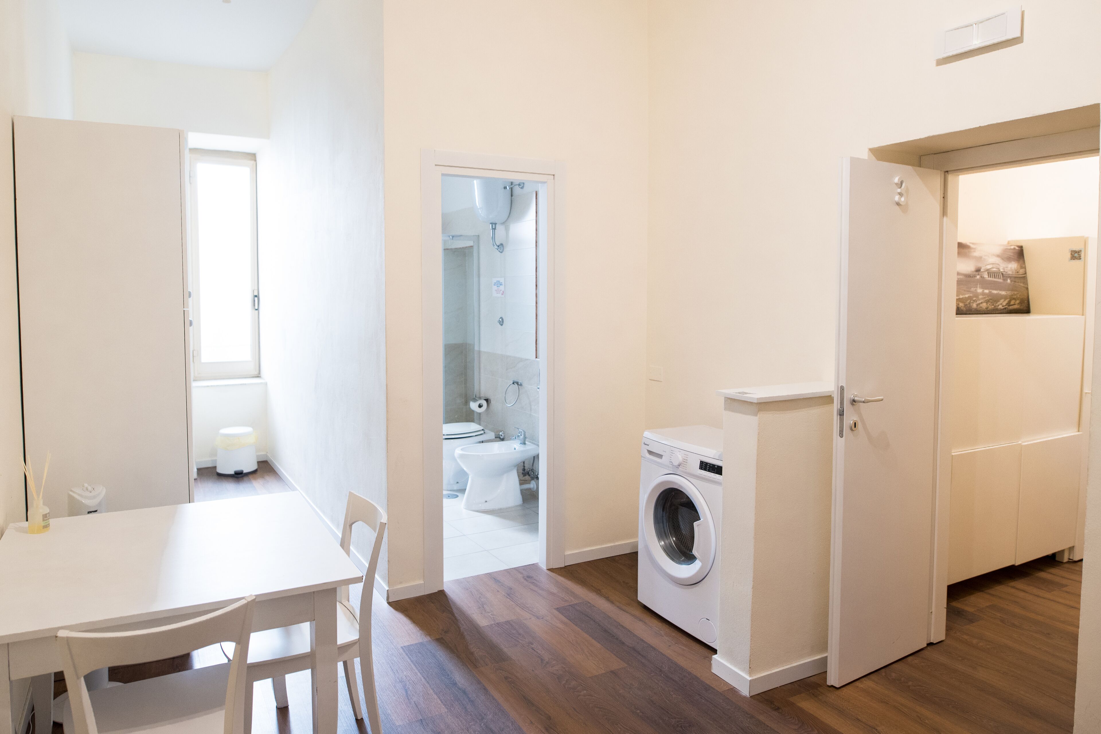 A compact kitchen with a washing machine, a dining area with a table and chairs, and a bathroom visible through an open door.