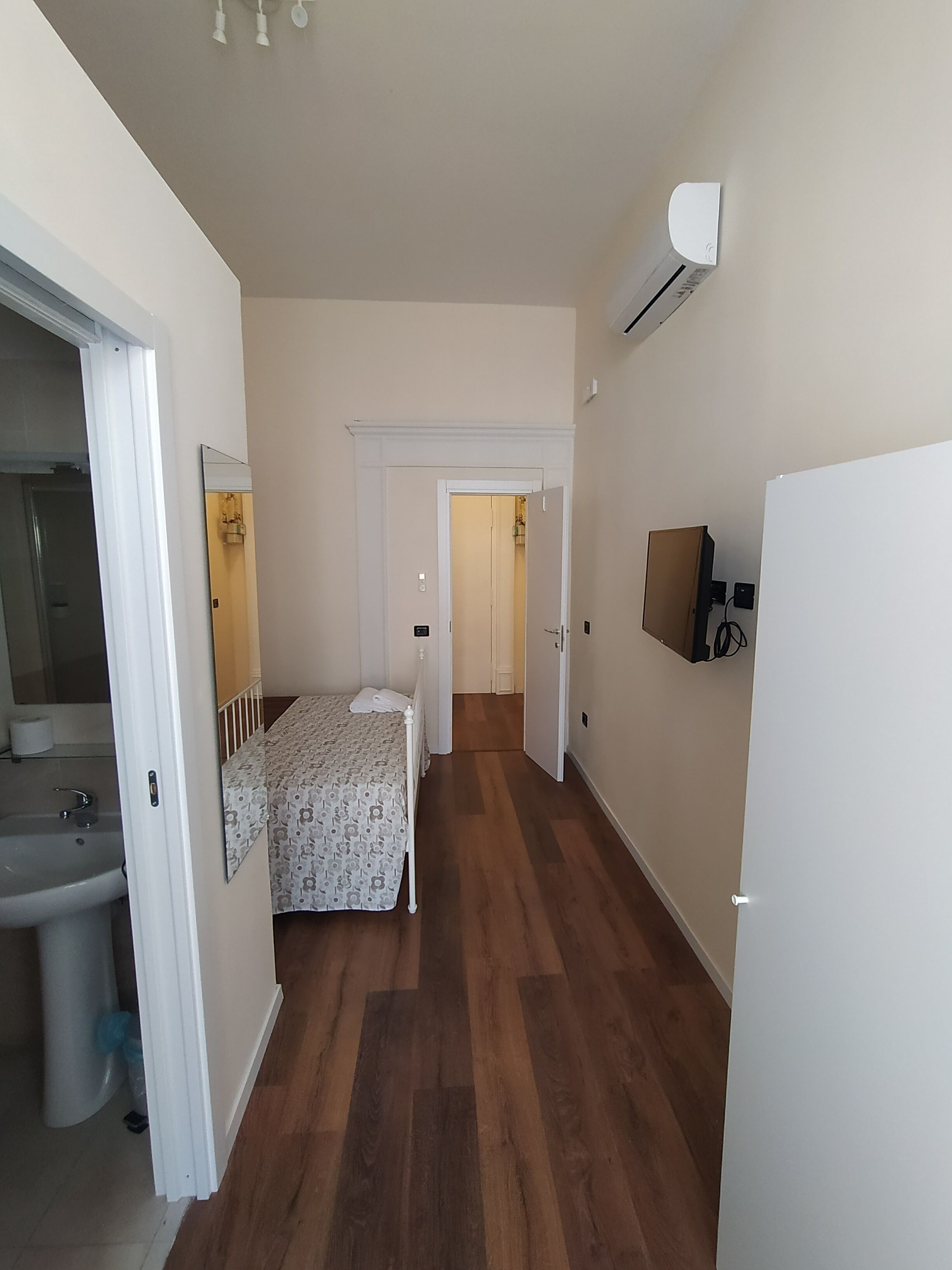 A narrow hallway with a bed, a sink, and a mirror on the left, and a television on the right.
