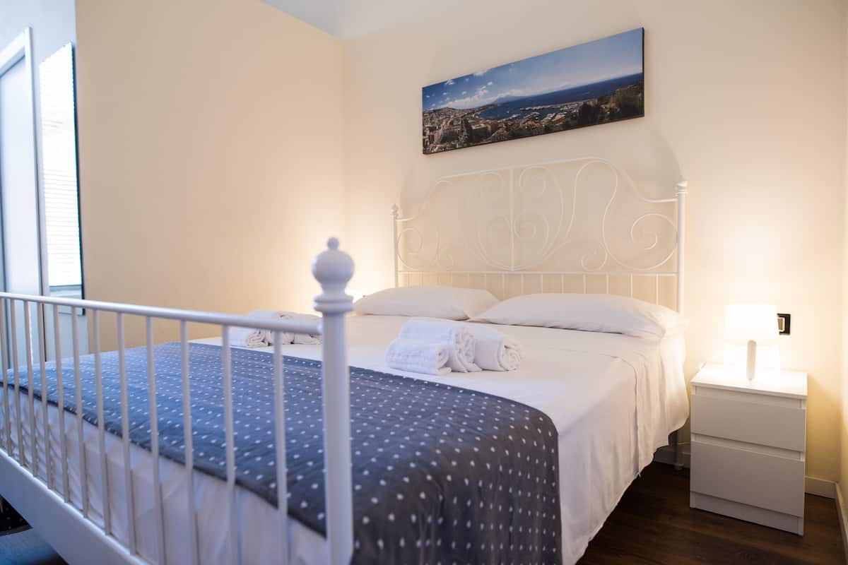 A neatly made bed with white linens and a dark polka dot bedspread, a white metal headboard, a bedside table with a lamp, and a cityscape painting on the wall.