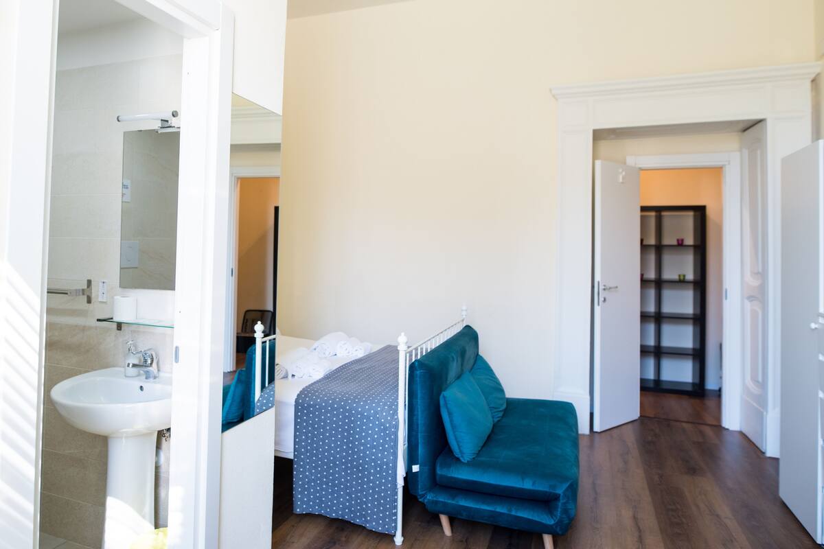 A bedroom with a bed, a bedside table, a window, and a bathroom with a sink and mirror.