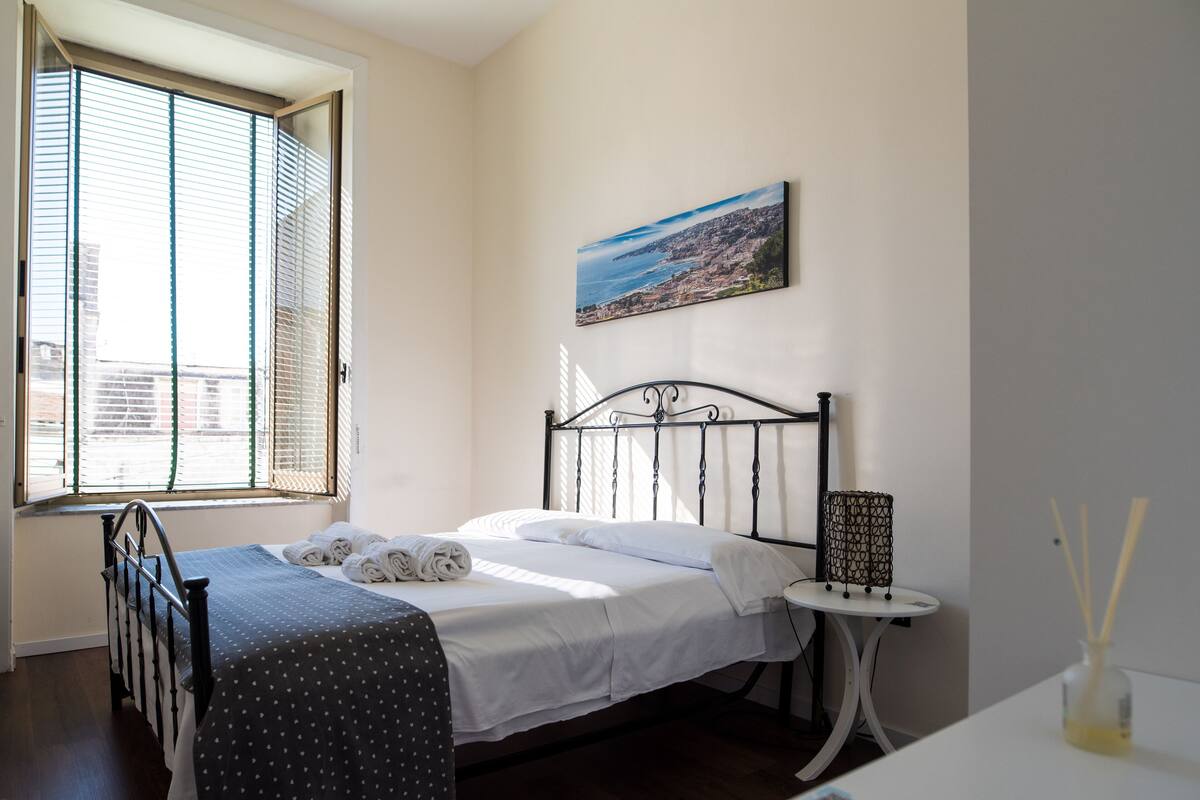 A neatly made bed with rolled towels, a small bedside table with a lamp, and a painting of a coastal landscape on the wall.