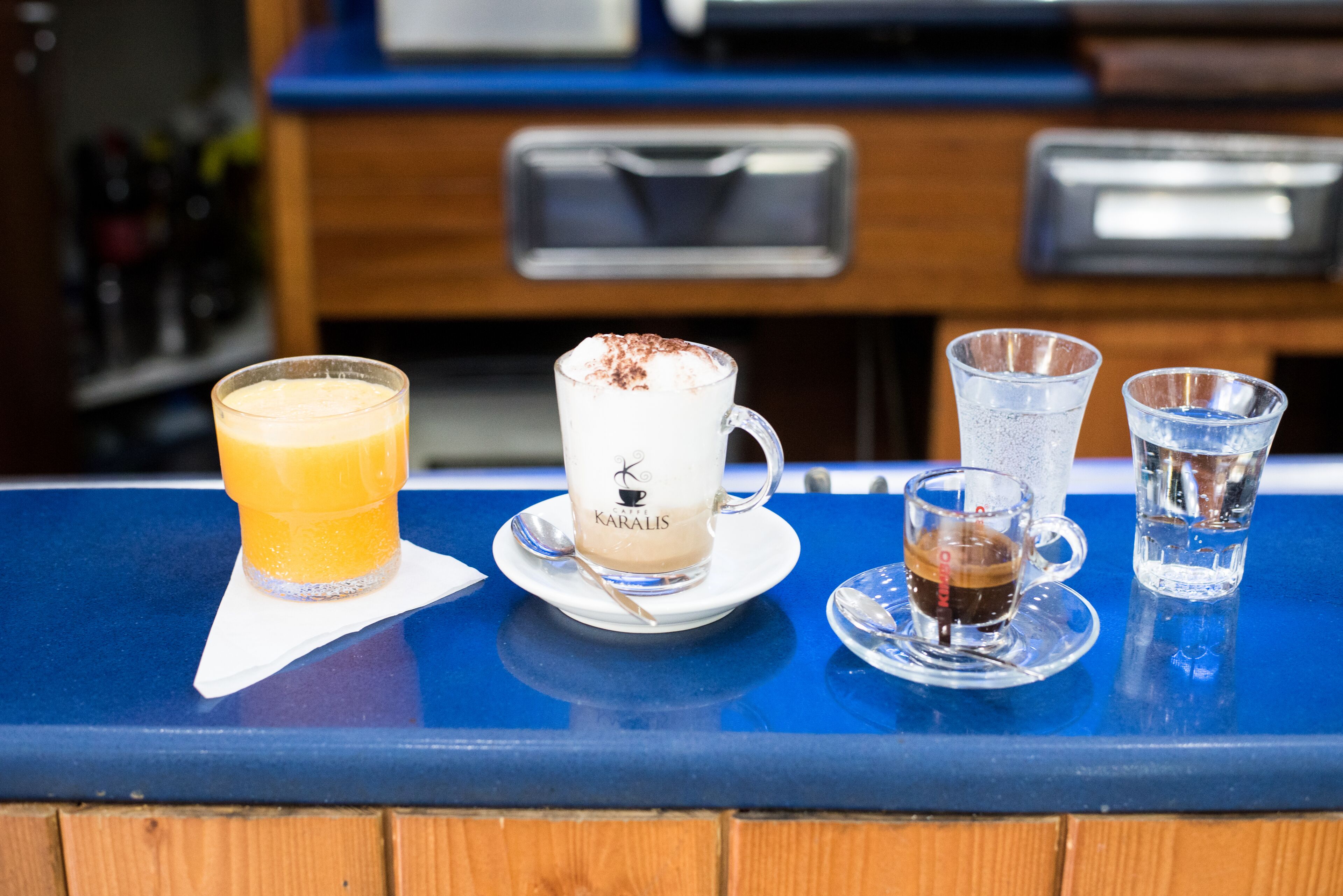 Una barra con diferentes bebidas, incluyendo un vaso de zumo de naranja, un capuchino y dos chupitos.