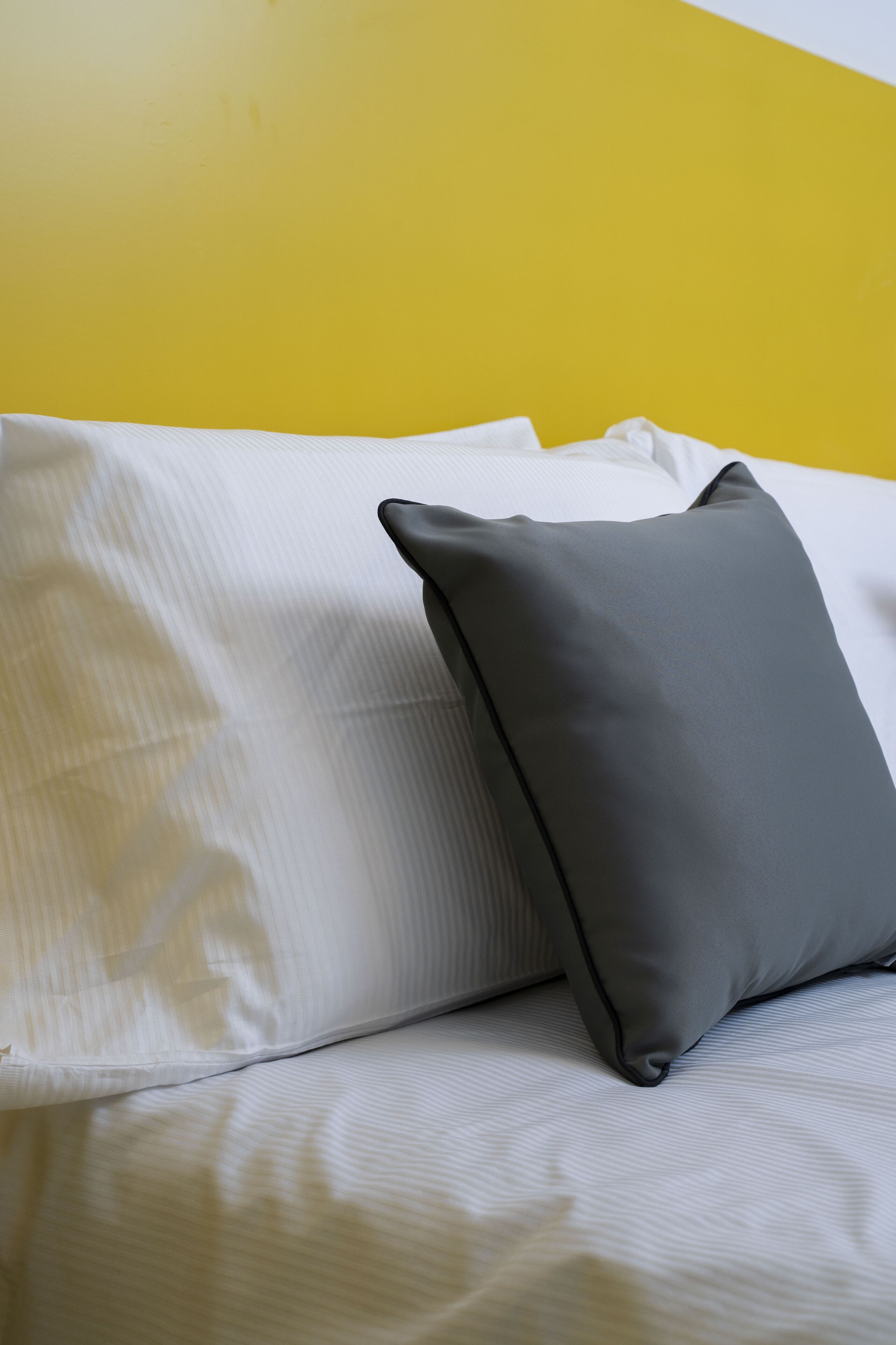 A bed with white bedding and a gray pillow against a yellow wall.