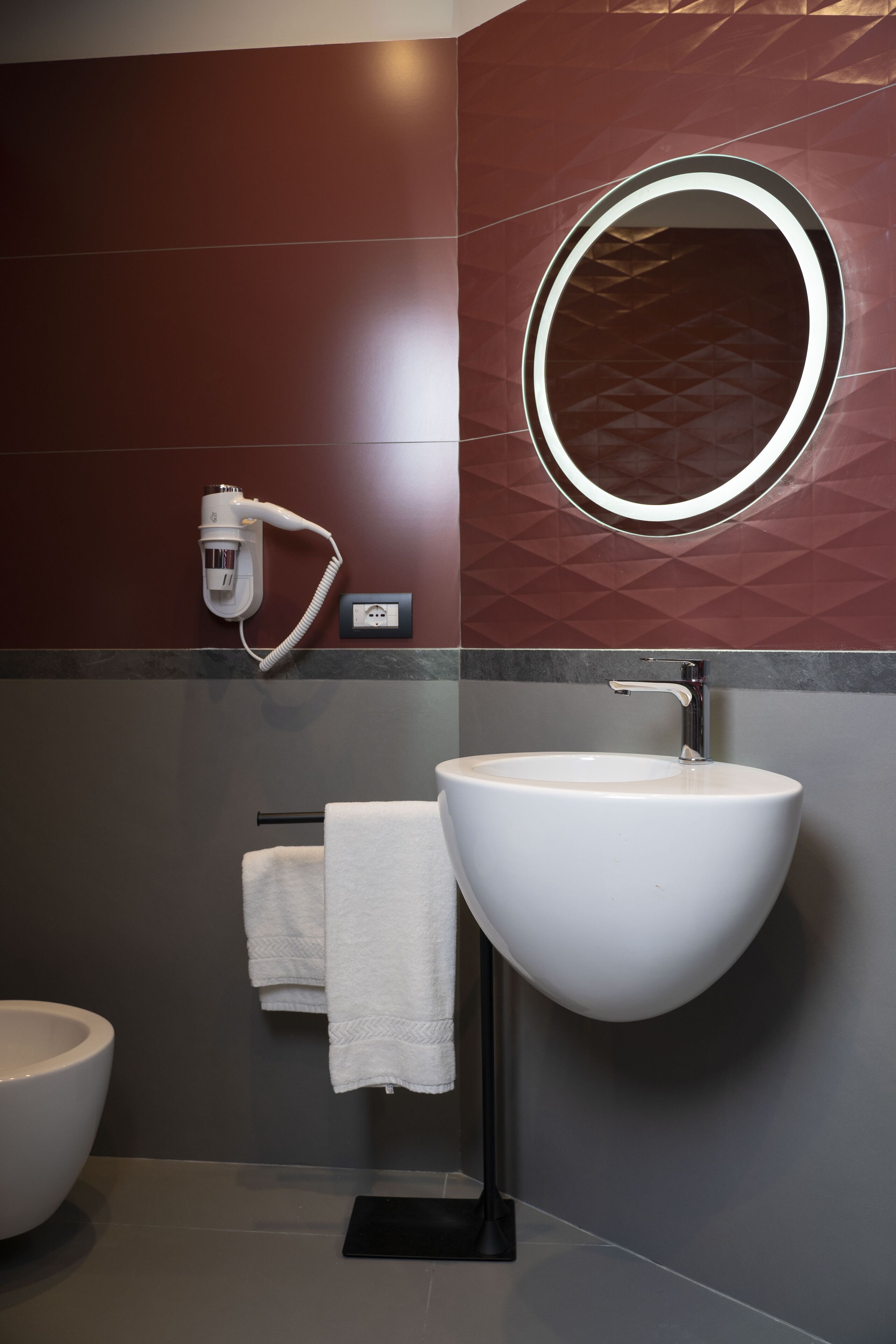 Un baño moderno con un lavamanos blanco, un espejo redondo y un secador de pelo montado en la pared.
