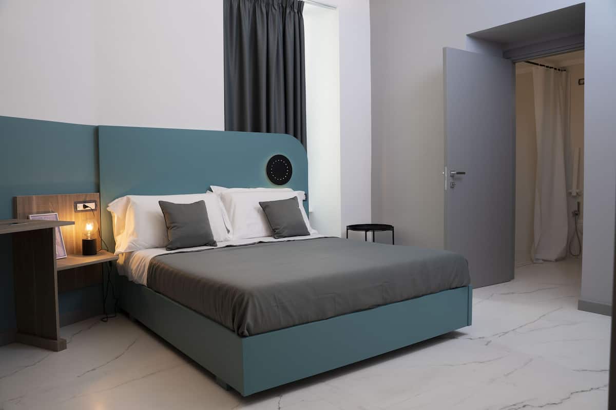 Une chambre moderne avec un lit de couleur turquoise, une table de chevet avec une lampe et une vue sur la salle de bain par une porte ouverte.