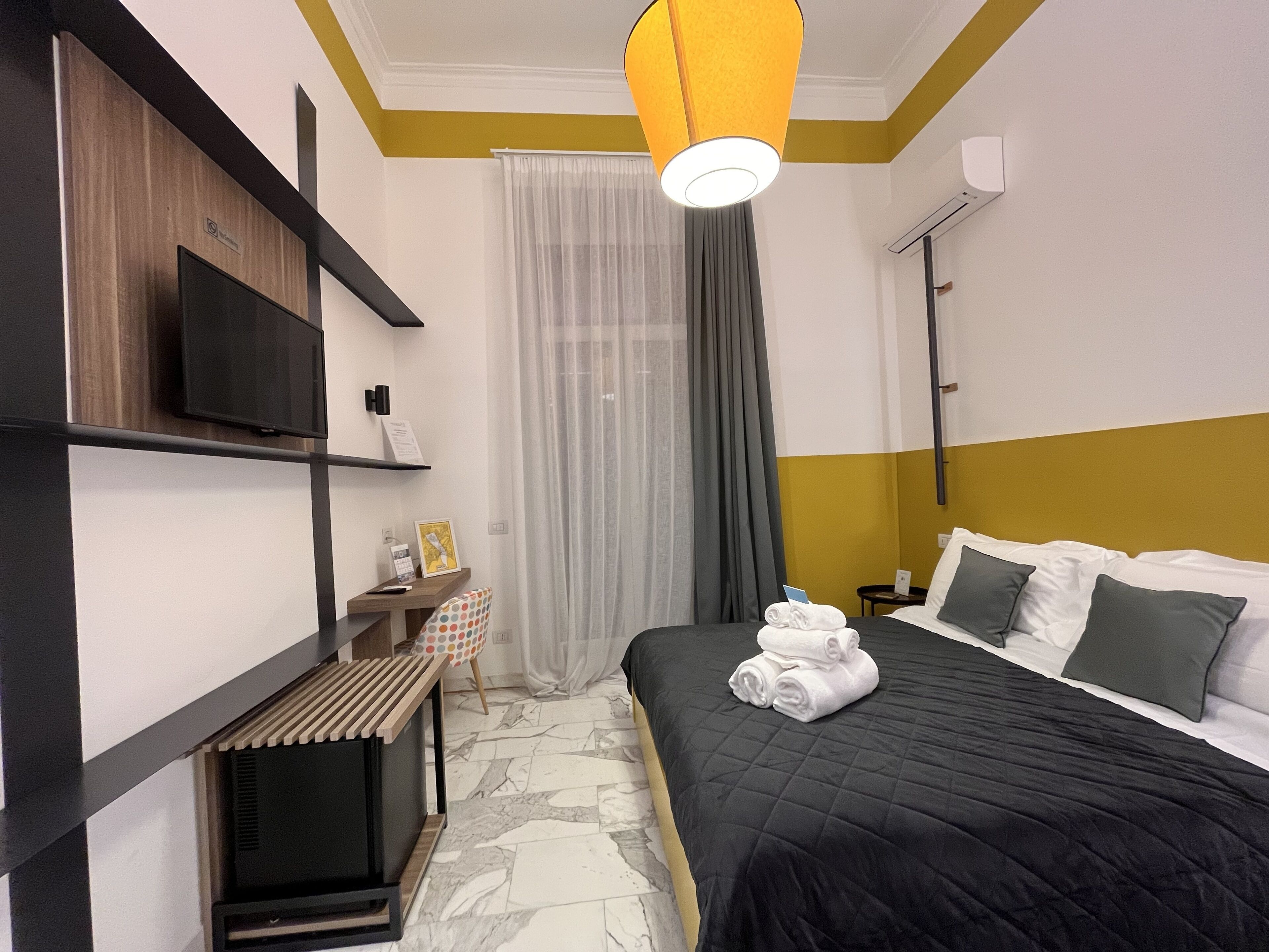 A hotel room with a bed, a TV mounted on the wall, a desk with a chair, and a window with curtains.
