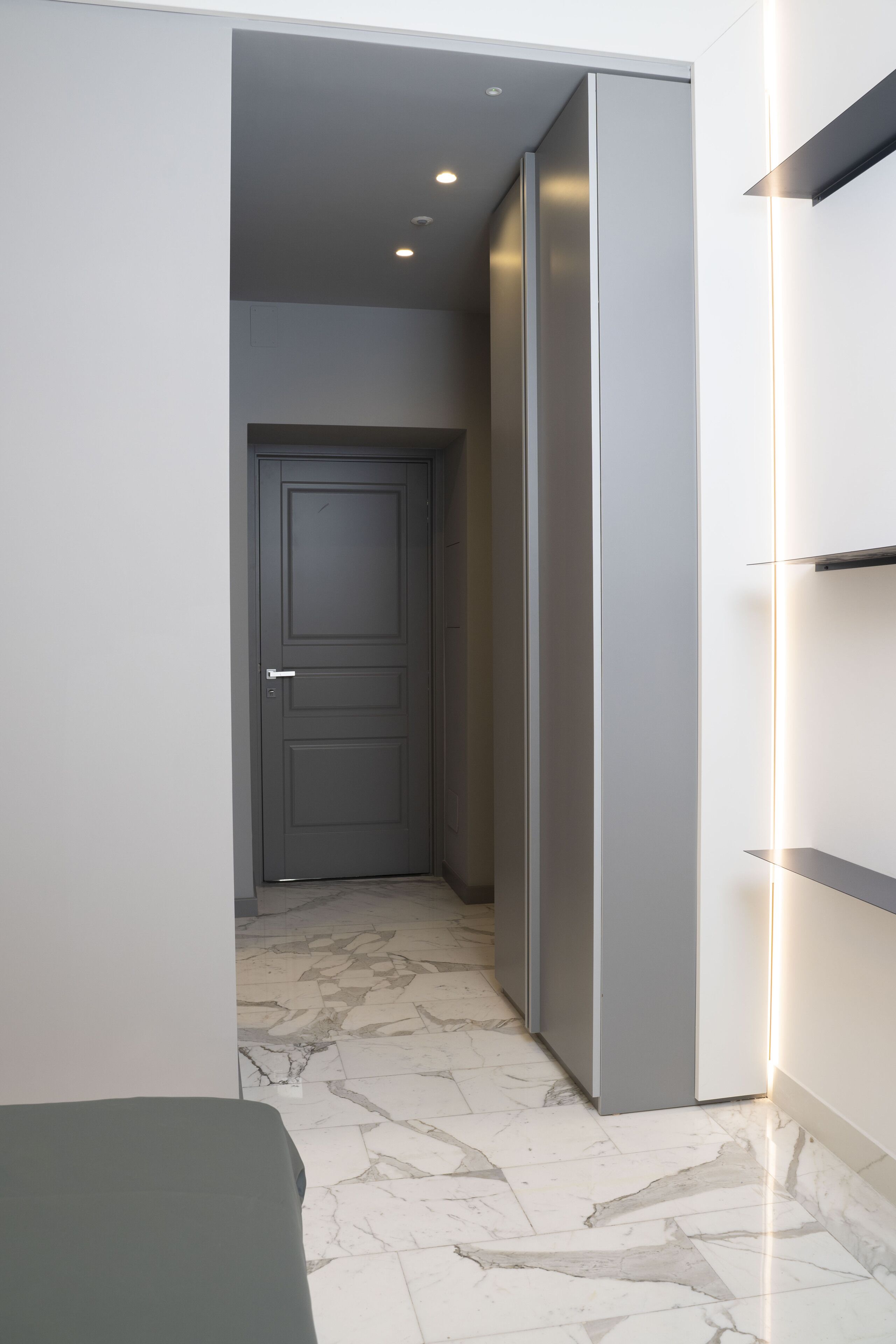 A modern hallway with a marble floor, a grey door, and a white wall with built-in shelves.