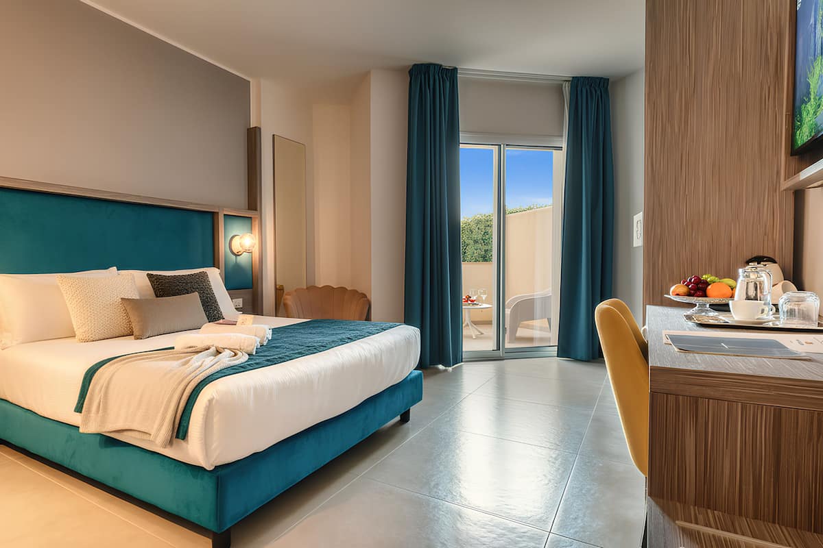 Ein Hotelzimmer mit Bett, Schreibtisch mit Stuhl, Esstisch mit Stühlen, einem Blumentopf und Blick ins Freie.