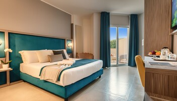 Habitación de hotel con cama, escritorio con silla, mesa de comedor con sillas, planta en maceta y vista al exterior.