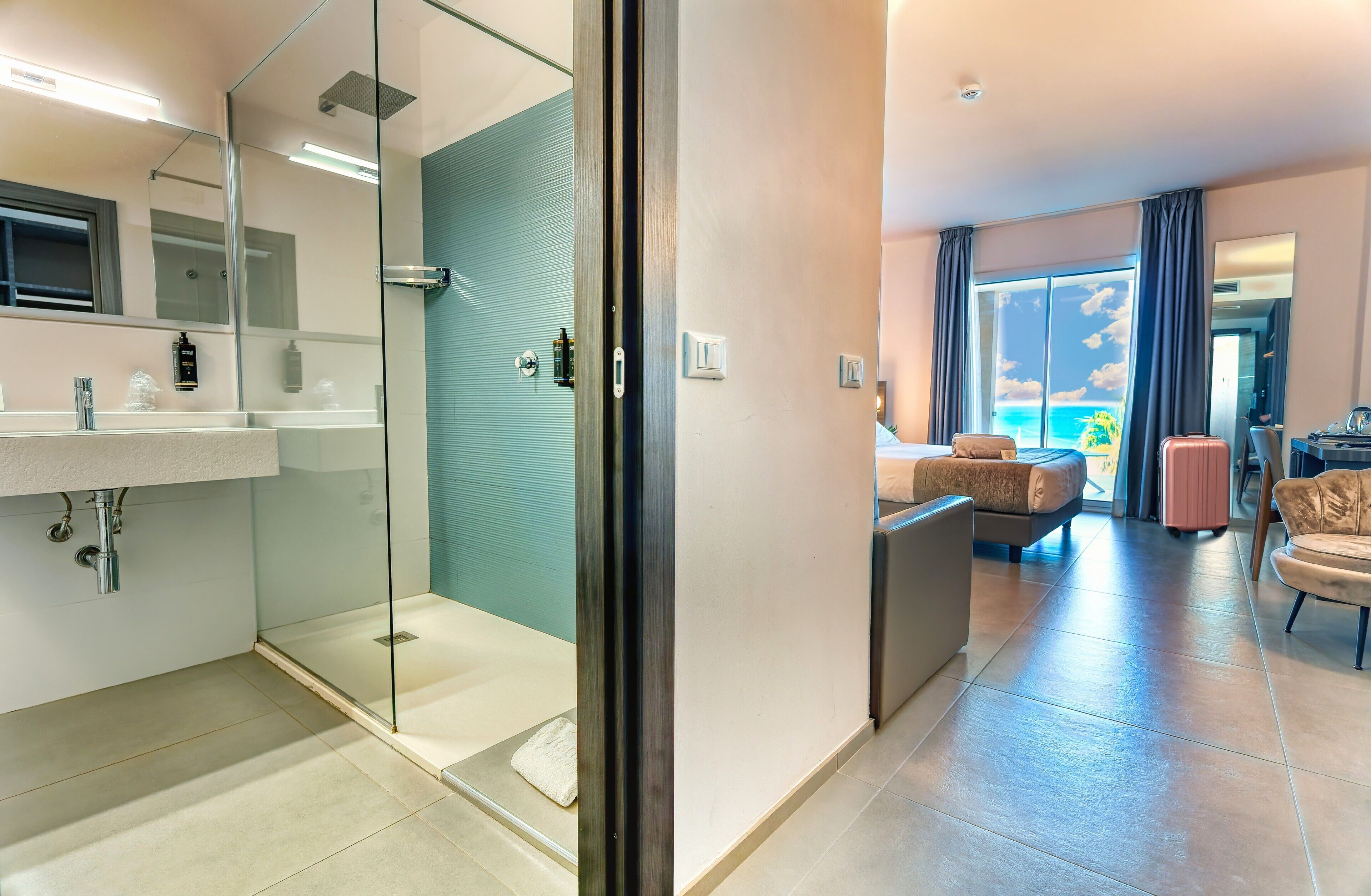 Una habitación de hotel moderna con ducha de vidrio, una cama grande, una zona de estar con vista al océano y una pequeña kitchenette.