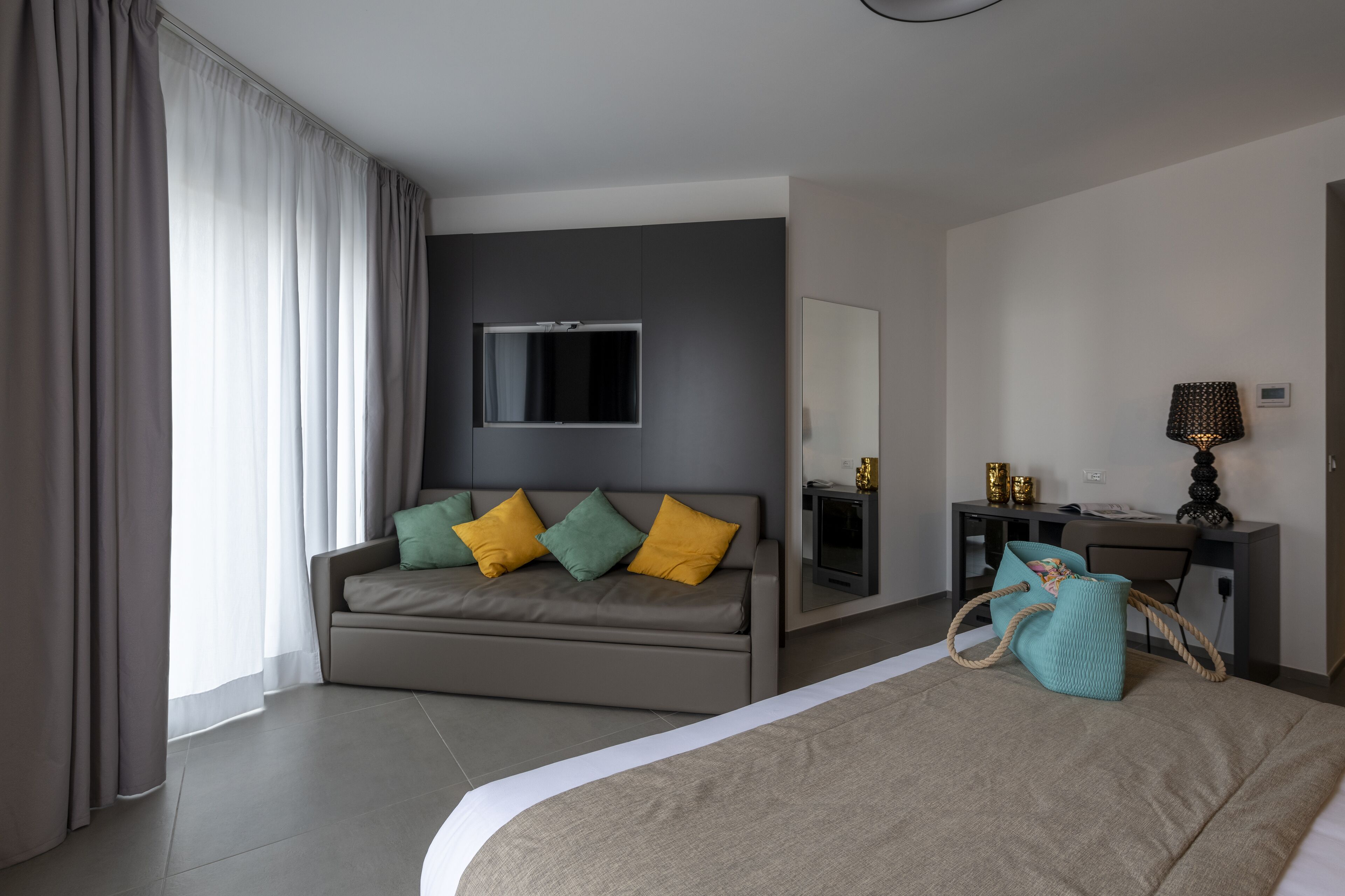 A modern hotel room with a grey sofa, a bed, a desk with a lamp, and a large window with curtains.
